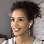 Mujer joven y sonriente con el cabello rizado recogido, auriculares inalámbricos y collar delicado, participando en una videollamada desde un salón con cojines y puertas de armario blancas al fondo, mirada cálida y expresión natural, 'PP afirma que la izquierda solo garantiza vivienda para okupas'.