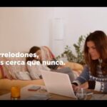 Mujer concentrada trabajando en un portátil en una mesa del salón mientras otra persona descansa en el sofá, con plantas, vela y objetos sobre la mesa en un ambiente hogareño y el titular visible PP de Torrelodones ofrece hasta 300 euros a propietarios con okupas.