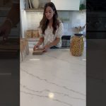 Una mujer está organizando varias bolsas de papel sobre una mesa de mármol, mientras a su lado hay un frasco grande lleno de palomitas de maíz, en un ambiente de cocina moderna, lo que refleja el evento de celebración donde 'Okupas en Granada organizan baby shower y generan escándalo'.