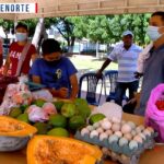 Vendedores y compradores con mascarillas atienden un puesto de mercado al aire libre con calabaza partida, aguacates, huevos en bandejas y bolsas con productos sobre una mesa bajo un toldo en un parque, en una imagen que refleja comunidad local y denuncia social: 'Okupas en Barajas enfrentan a vecinos por amenazas de muerte'.