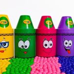 Filas de recipientes con forma de crayón en rojo, amarillo, verde, rosa, morado y azul con caras expresivas sobre una superficie cubierta de bolitas multicolores, Vecinos de Casas de Colores en Campclar exigen dignidad residencial.