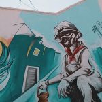 Mural vibrante que combina un retrato de un hombre con un diseño de un personaje sentado, vistiendo overol y con una máscara, rodeado de elementos urbanos, reflejando un mensaje de resistencia y arte social en el contexto de la 'Revuelta ciudadana contra la okupación en Albiol y vecinos'.