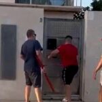 Imagen diurna de tres hombres junto a una puerta metálica en la fachada de una vivienda, uno usando una palanca roja intentando forzar la cerradura mientras los otros observan junto a un coche, ilustrando Cómo expulsar rápidamente a okupas de viviendas en España.