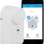 Unidad de cierre de puerta inteligente blanca mostrada en primer plano junto a un teléfono móvil que muestra la app de control, eqiva Unidad de cierre de puerta inteligente Bluetooth, blanca, 142950A0, 5,6 x 5,2 x 11,4 cm, ideal para control remoto, cierre automático y fácil instalación.