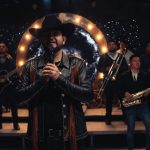 Cantante principal con sombrero vaquero y chaqueta de cuero con flecos sostiene un micrófono mientras detrás de él una banda con guitarras, acordeón, saxofón y trombones actúa frente a un aro de luces circular y un fondo estrellado, Okupas amenazan de muerte en Granada tras siete años de ocupación