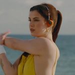 Mujer joven con coleta y pendientes dorados estira los brazos frente al mar con auriculares naranja y vestido amarillo en una playa soleada, con tatuaje en el antebrazo y el logo de Vevo parcialmente visible; Justicia ordena desalojo del Hotel Callao Sport en Tenerife.
