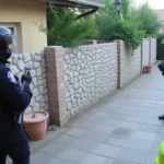 Dos agentes con chalecos y cascos protectores sosteniendo un arma larga y una persona con chaleco reflectante vigilan un pasillo peatonal junto a un muro de piedra frente a una vivienda, Increíble enfrentamiento: dos okupas intentan aferrarse a una casa y desafían a la Guardia Civil en una increíble resistencia