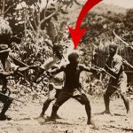 Fotografía en sepia que muestra a un grupo de hombres con lanzas y palos rodeando a una persona de pie con los brazos abiertos en un entorno de chozas y vegetación, indicada por una flecha roja, Okupa debe 16.000 euros y amenaza a propietaria con destrozar piso.