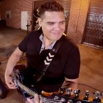 Guitarrista sonriente tocando una guitarra eléctrica en primer plano junto a otro músico con gorra y un micrófono en un escenario al aire libre frente a un muro de ladrillo, imagen de concierto nocturno que incluye el titular "Pesadilla en Alcorcón por okupas: robos, peleas y drogas".