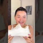 Mujer joven sonriente en interior sosteniendo una zapatilla deportiva blanca frente a la cámara, con uñas pintadas de lila y un cuadro en la pared de fondo; La Justicia anula desahucio por falta de negociación del propietario