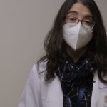 Mujer profesional de la salud con bata blanca, gafas y mascarilla blanca, cabello suelto y bufanda oscura, con placa identificativa y gesto con la mano frente a un fondo neutro, La alcaldesa de Alcorcón enfrenta problemas de convivencia por okupas