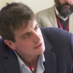 Plano medio de un hombre joven vestido con americana y camisa hablando al micrófono en una reunión interior mientras, al fondo, otro hombre con barba y chaqueta clara escucha atento; Dos inquilinos residen en una vivienda de Portugalete