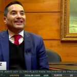 Hombre con traje azul y corbata roja sentado en una sala con paneles de madera y un cuadro enmarcado durante una entrevista televisiva con logo de noticias, recuadro de intérprete de lengua de señas y tira informativa en la parte inferior, Viviendas en venta en Gerona y Tarragona ocupadas.