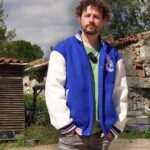 Retrato accesible de un hombre joven de pelo rizado y barba, con chaqueta azul y blanca tipo varsity y camiseta verde, de pie con las manos en los bolsillos frente a un patio rural con muros de piedra, cobertizo de madera y escombros que sugieren tensión en la vivienda; Inquiokupa estalla contra su casera y defiende su posición
