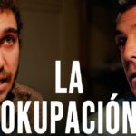 Primer plano de dos hombres en diálogo serio con iluminación cálida y el gran texto blanco "LA OKUPACIÓN" en el centro, imagen enfocada en expresiones faciales y tema de vivienda, Andalucía critica a Pedro Sánchez por favorecer a okupas
