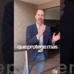 Un hombre de pie en un pasillo exterior, con chaqueta azul y vaqueros, gesticulando con expresión entusiasta frente a una barandilla y una pared de piedra, con texto superpuesto 'que protege más' y un pequeño logotipo en la esquina, Descubre por qué desalojaron a unos okupas de una casa en el Mediterráneo en tiempo récord: la impactante confesión del propietario te dejará sin palabras.