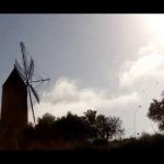 Silueta en contraluz de un molino tradicional sobre una loma con arbustos y árboles bajo un cielo brillante y soleado, imagen ilustrativa con el titular 'Vallan un edificio en Mallorca para evitar okupación'.