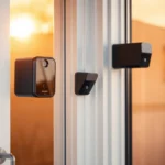 Sensores anti-okupas: puertas y ventanas, dispositivos negros compactos tipo sensor y cámara montados en el marco de una puerta corredera de cristal al atardecer para vigilancia y detección de intrusiones.