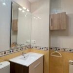 Baño pequeño y luminoso con lavabo moderno sobre mueble de madera, inodoro, amplio espejo sobre encimera, azulejos claros con cenefa decorativa, mampara de bloques de vidrio, armario alto y radiador, Okupas en la urbanización de Arroyo se benefician de la Sareb.