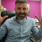 Hombre con barba y cabello corto, sentado en una silla de oficina, gesticulando con las manos mientras habla, en un entorno de oficina con paredes de color rosa y muebles de madera, recuperan vivienda ocupada en Fisterra tras más de un año.